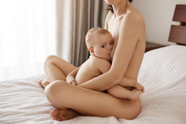 Young attractive nude mom breastfeeding hugging her newborn baby smiling sitting on bed at home.