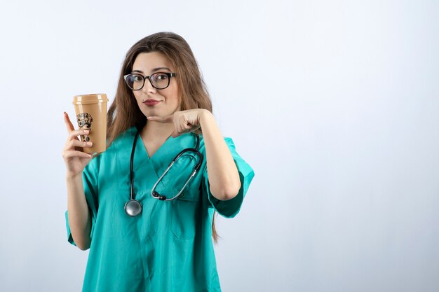 Young attractive nurse with stethoscope pointing at a cup of coffee 