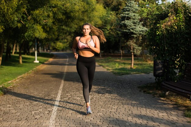 ピンクのスポーティなトップとレギンスの若い魅力的なプラスサイズの女性は、都市公園で走っている間、カメラで思慮深く見ています