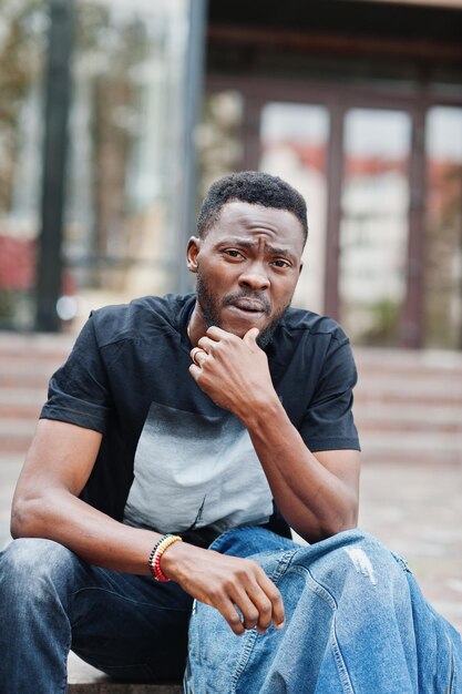 Young attractive serious african american man model Modern handsome guy in jeans jacket sitting on urban city street