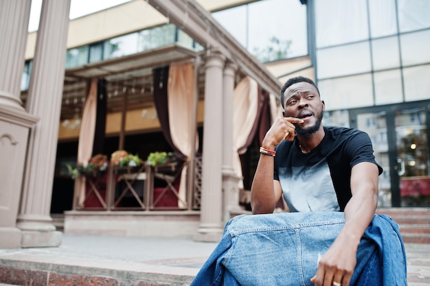 Free photo young attractive serious african american man model modern handsome guy in jeans jacket sitting on urban city street