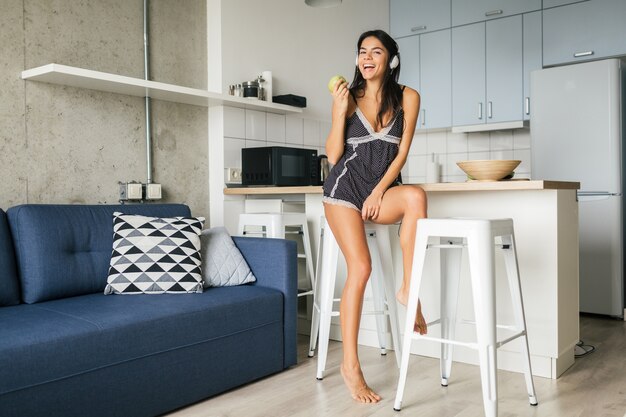 Young attractive sexy woman having breakfast in stylish modern kitchen in morning, eating apple, smiling, happy, positive, healthy lifestyle, listening to music on headphones, laughing, having fun