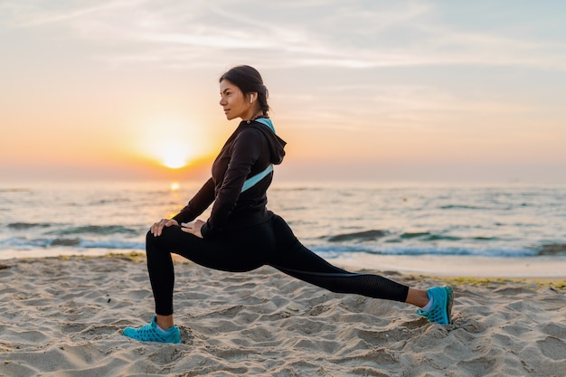 Young attractive slim woman doing sport exercises on morning sunrise beach in sports wear, healthy lifestyle, listening to music on earphones, making stretching for legs