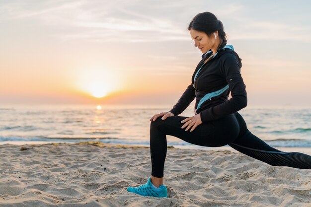 Young attractive slim woman doing sport exercises on morning sunrise beach in sports wear, healthy lifestyle, listening to music on earphones, making stretching for legs