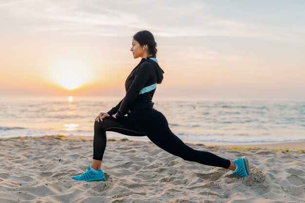 Young attractive slim woman doing sport exercises on morning sunrise beach in sports wear, healthy lifestyle, listening to music on earphones, making stretching for legs