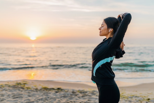 Young attractive slim woman doing sport exercises on morning sunrise beach in sports wear, healthy lifestyle, listening to music on earphones, making stretching
