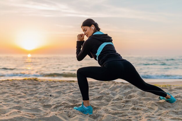 Young attractive slim woman doing sport exercises on morning sunrise beach in sports wear, healthy lifestyle, listening to music on earphones, making stretching