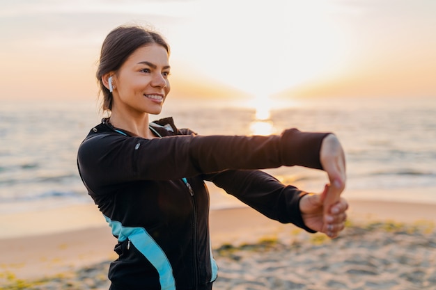 Молодая привлекательная стройная женщина делает спортивные упражнения на утреннем пляже восхода солнца в спортивной одежде, здоровом образе жизни, слушает музыку в наушниках, делает растяжку
