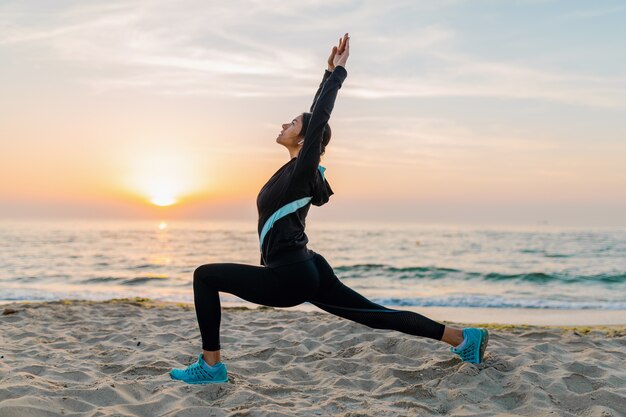 Молодая привлекательная стройная женщина делает спортивные упражнения на утреннем пляже восхода солнца в спортивной одежде, здоровом образе жизни, слушает музыку в наушниках, занимается йогой
