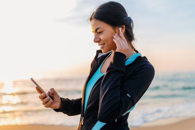 Young attractive slim woman doing sport exercises on morning sunrise beach in sports wear, healthy lifestyle, listening to music on wireless earphones holding smartphone, smiling happy having fun