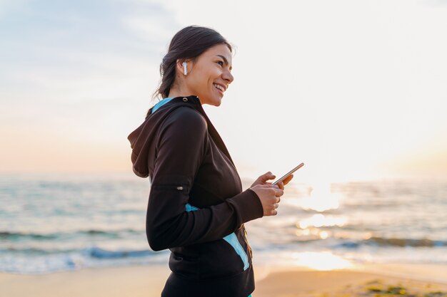 Young attractive slim woman doing sport exercises on morning sunrise beach in sports wear, healthy lifestyle, listening to music on wireless earphones holding smartphone, smiling happy having fun