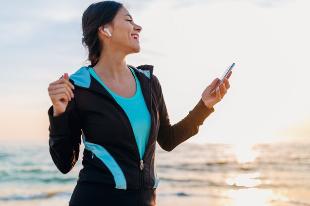 Young attractive slim woman doing sport exercises on morning sunrise beach in sports wear, healthy lifestyle, listening to music on wireless earphones holding smartphone, smiling happy having fun