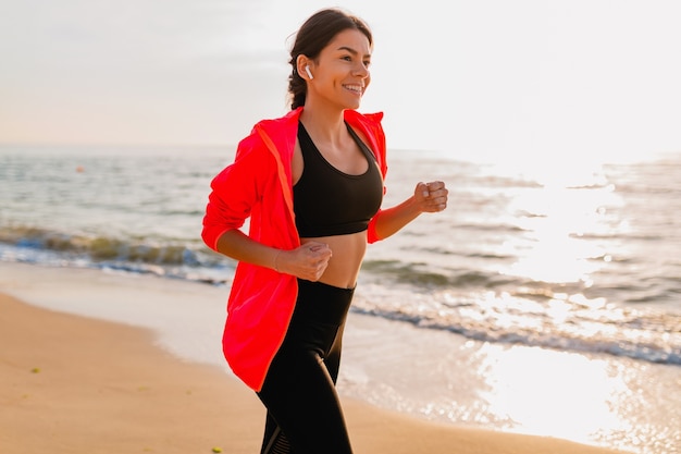 Молодая привлекательная стройная женщина делает спортивные упражнения на утреннем восходе солнца, бегая трусцой на морском пляже в спортивной одежде, здоровом образе жизни, слушая музыку в наушниках, в розовой ветровке