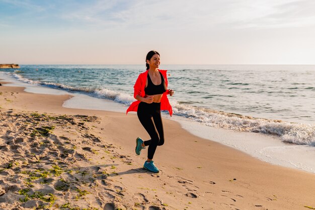Молодая привлекательная стройная женщина делает спортивные упражнения на утреннем восходе солнца, бегая трусцой на морском пляже в спортивной одежде, здоровом образе жизни, слушая музыку в наушниках, в розовой ветровке