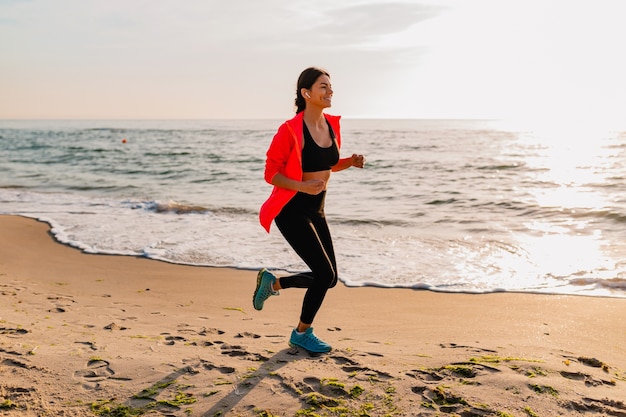 Young attractive slim woman doing sport exercises in morning sunrise jogging on sea beach in sports wear, healthy lifestyle, listening to music on earphones, wearing pink windbreaker jacket