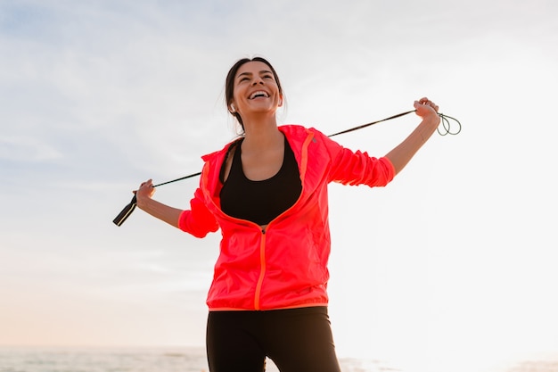 Young attractive slim woman doing sport exercises in morning sunrise on sea beach in sports wear, healthy lifestyle, listening to music on earphones, wearing pink windbreaker jacket, holding jump rope