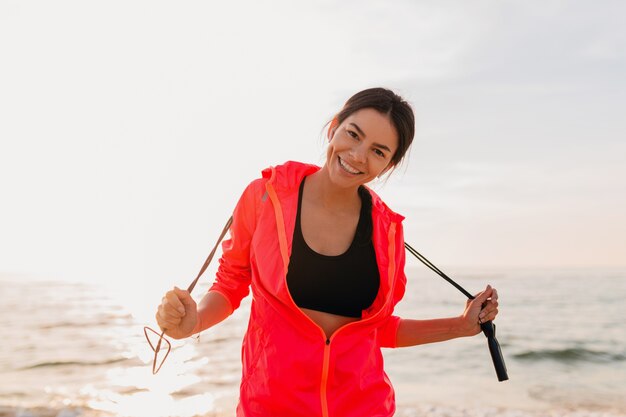 Young attractive slim woman doing sport exercises in morning sunrise on sea beach in sports wear, healthy lifestyle, wearing pink windbreaker jacket, holding jump rope