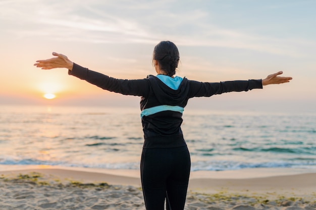 Young attractive slim woman greeting sun ciew from back holding hands doing sport exercises on morning sunrise beach in sports wear, healthy lifestyle