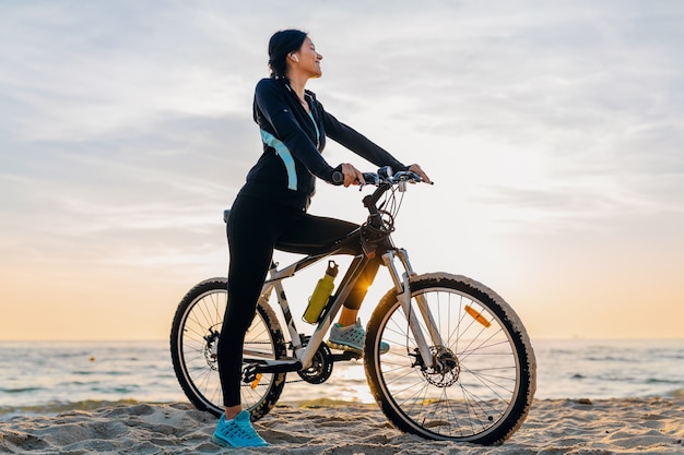 Young attractive slim woman riding bicycle, sport in morning sunrise summer beach in sports fitness wear, active healthy lifestyle, smiling happy having fun