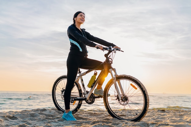 Young attractive slim woman riding bicycle, sport on morning sunrise summer beach in sports fitness wear, active healthy lifestyle, smiling happy having fun