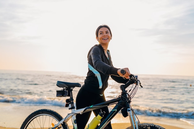 Young attractive slim woman riding bicycle, sport in morning sunrise summer beach in sports fitness wear, active healthy lifestyle, smiling happy having fun