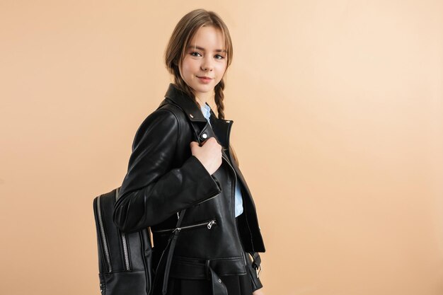 Young attractive smiling girl with two braids in leather jacket and skirt with black backpack on shoulder while happily looking in camera over beige background