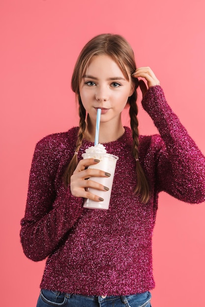 Молодая привлекательная улыбающаяся девушка с двумя косами в свитере держит в руках молочный коктейль, мечтательно глядя в камеру на розовом фоне