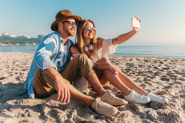 Young attractive smiling happy man and woman in sunglasses sitting on sand beach taking selfie photo