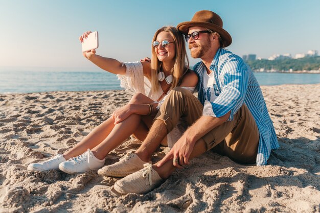 Молодой привлекательный улыбающийся счастливый мужчина и женщина в солнцезащитных очках, сидя на песчаном пляже, делая селфи фото
