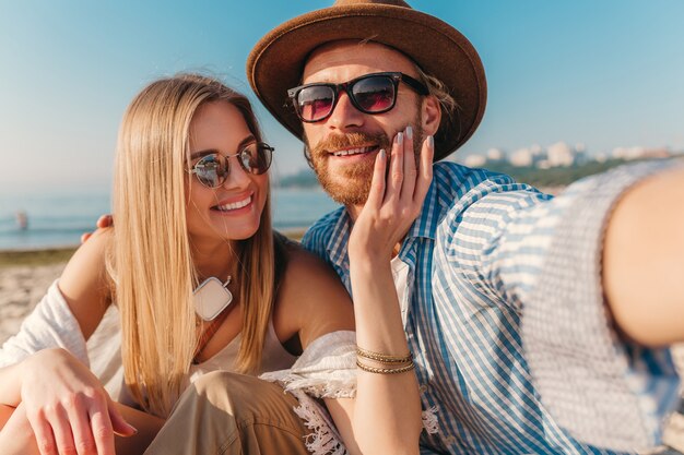 Young attractive smiling happy man and woman in sunglasses sitting on sand beach taking selfie photo