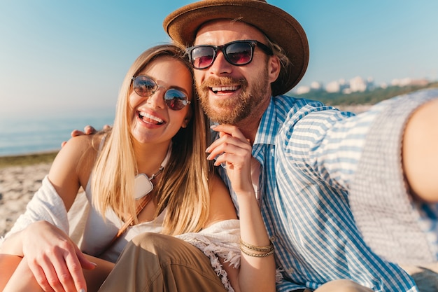 Young attractive smiling happy man and woman in sunglasses sitting on sand beach taking selfie photo