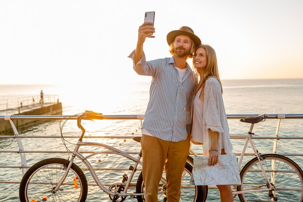 젊은 매력적인 웃는 행복 한 남자와 여자는 휴대 전화 카메라에 selfie 사진을 복용 자전거 여행, 일몰에 바다로 로맨틱 커플, boho 힙 스터 스타일의 옷, 친구가 함께 재미