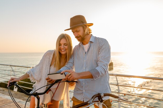 若い魅力的な笑顔幸せな男とスマートフォンを使用して自転車で旅行する女性、夕日に海でロマンチックなカップル、自由奔放に生きるヒップスタイルの服、一緒に楽しんで友達