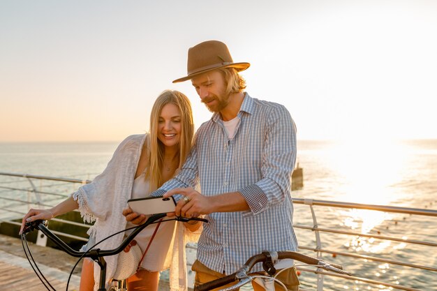 젊은 매력적인 웃는 행복 한 남자와 여자는 스마트 폰을 사용하여 자전거 여행, 일몰에 바다로 로맨틱 커플, boho 힙 스터 스타일의 옷, 친구가 함께 재미