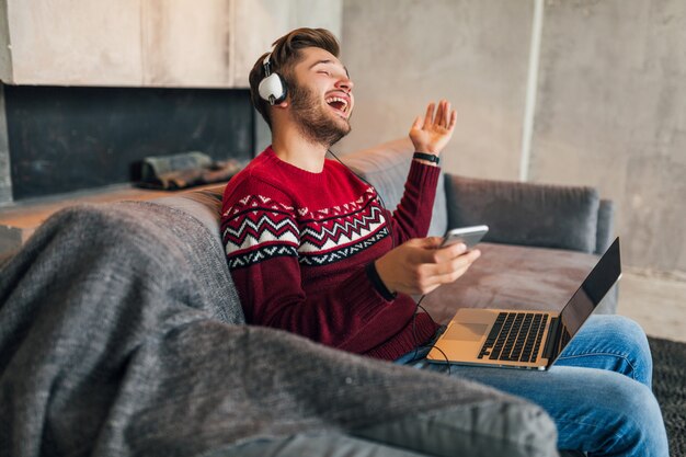 Молодой привлекательный улыбающийся мужчина на диване у себя дома зимой поет под музыку в наушниках, в красном вязаном свитере, работает на ноутбуке, фрилансер, эмоциональный, смеющийся, счастливый