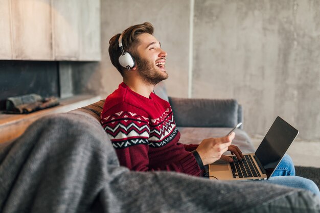 Young attractive smiling man on sofa at home in winter singing to music on headphones, wearing red knitted sweater, working on laptop, freelancer, emotional, laughing, happy