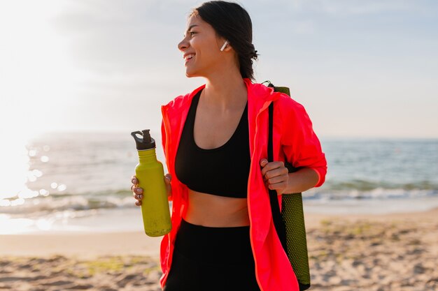 Young attractive smiling woman doing sports in morning sunrise on sea beach holding yoga mat and bottle of water, healthy lifestyle, listening to music on earphones, wearing pink windbreaker jacket