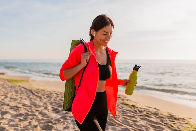 Young attractive smiling woman doing sports in morning sunrise on sea beach holding yoga mat and bottle of water, healthy lifestyle, listening to music on earphones, wearing pink windbreaker jacket