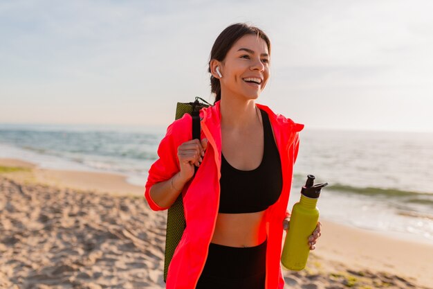 Young attractive smiling woman doing sports in morning sunrise on sea beach holding yoga mat and bottle of water, healthy lifestyle, listening to music on earphones, wearing pink windbreaker jacket