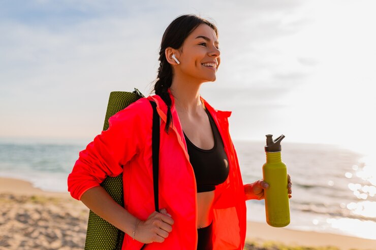 Free Photo young attractive smiling woman doing sports in morning sunrise on sea beach holding yoga mat and bottle of water, healthy lifestyle, listening to music on earphones, wearing pink windbreaker jacket