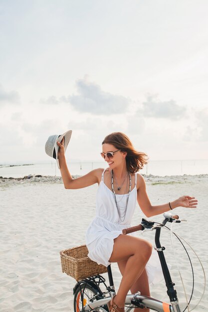 Young attractive smiling woman in white dress riding on tropical beach on bicycle wearing hat and sunglasses