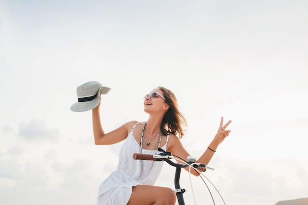 Young attractive smiling woman in white dress riding on tropical beach on bicycle wearing hat and sunglasses