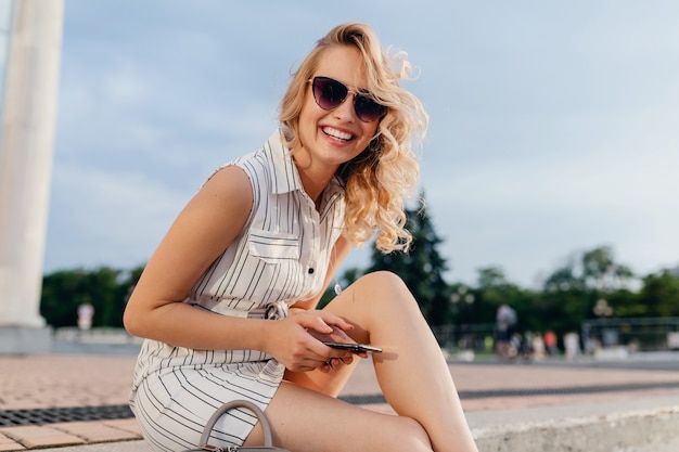 Young attractive stylish blonde woman sitting in city street in summer fashion style dress wearing sunglasses, holding phone, laughing candid