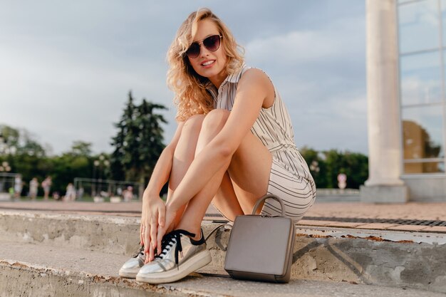 Young attractive stylish blonde woman sitting in city street in summer fashion style dress wearing sunglasses, purse, silvers sneakers 