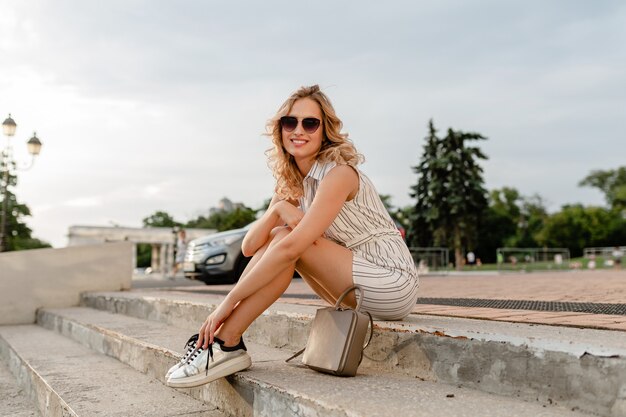 Young attractive stylish blonde woman sitting in city street in summer fashion style dress wearing sunglasses, purse, silvers sneakers 