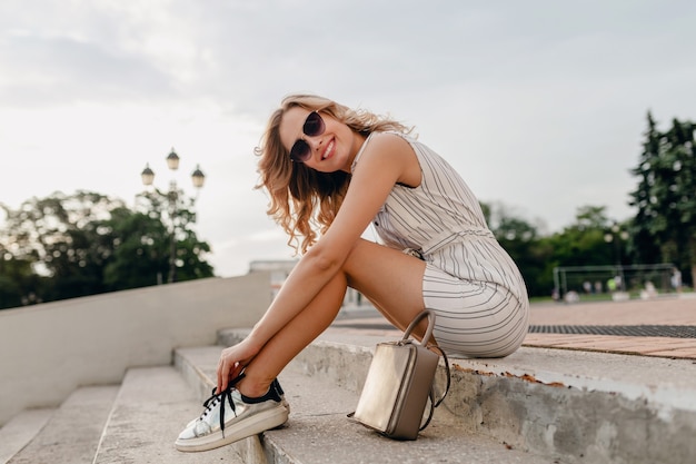 サングラス、財布、銀のスニーカーを身に着けている夏のファッションスタイルのドレスで街の通りに座っている若い魅力的なスタイリッシュなブロンドの女性