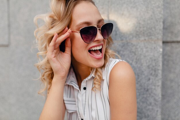 Young attractive stylish blonde woman walking in city street in summer fashion style dress wearing sunglasses, laughing surprised