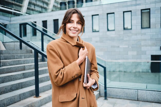 Young attractive stylish businesswoman in coat with laptop dreamily looking away outdoor