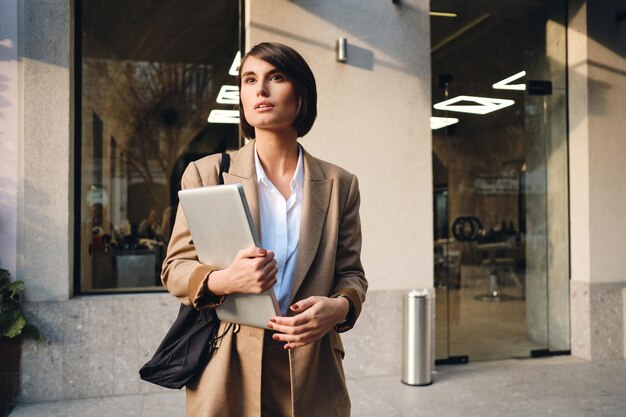 Young attractive stylish businesswoman with laptop dreamily looking away on city street