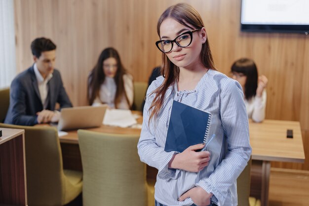 Young attractive stylish office worker girl in glasses with a notebook in the hands on background of working colleagues